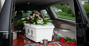 Carro fúnebre transportando caixão branco com flores brancas em cima