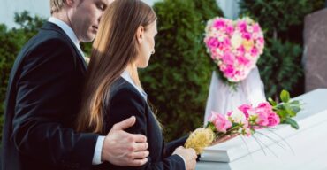 Uma mulher, vestindo preto, está ao lado de um caixão branco com flores. Um homem a conforta com a mão no ombro, transmitindo a emoção de um discurso fúnebre.