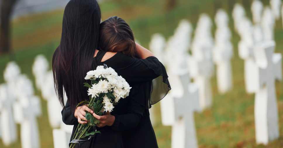 Duas mulheres abraçam-se num cemitério enquanto seguram flores, em meio ao suporte no luto familiar.